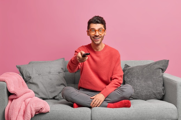 People home lifestyle leisure concept. Cheerful relaxed guy sits in lotus pose on comfortable sofa holds remote control