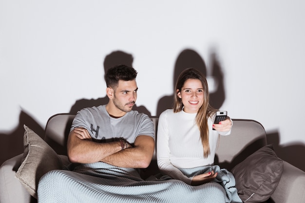 Smiling woman changing TV channels near angry man on sofa